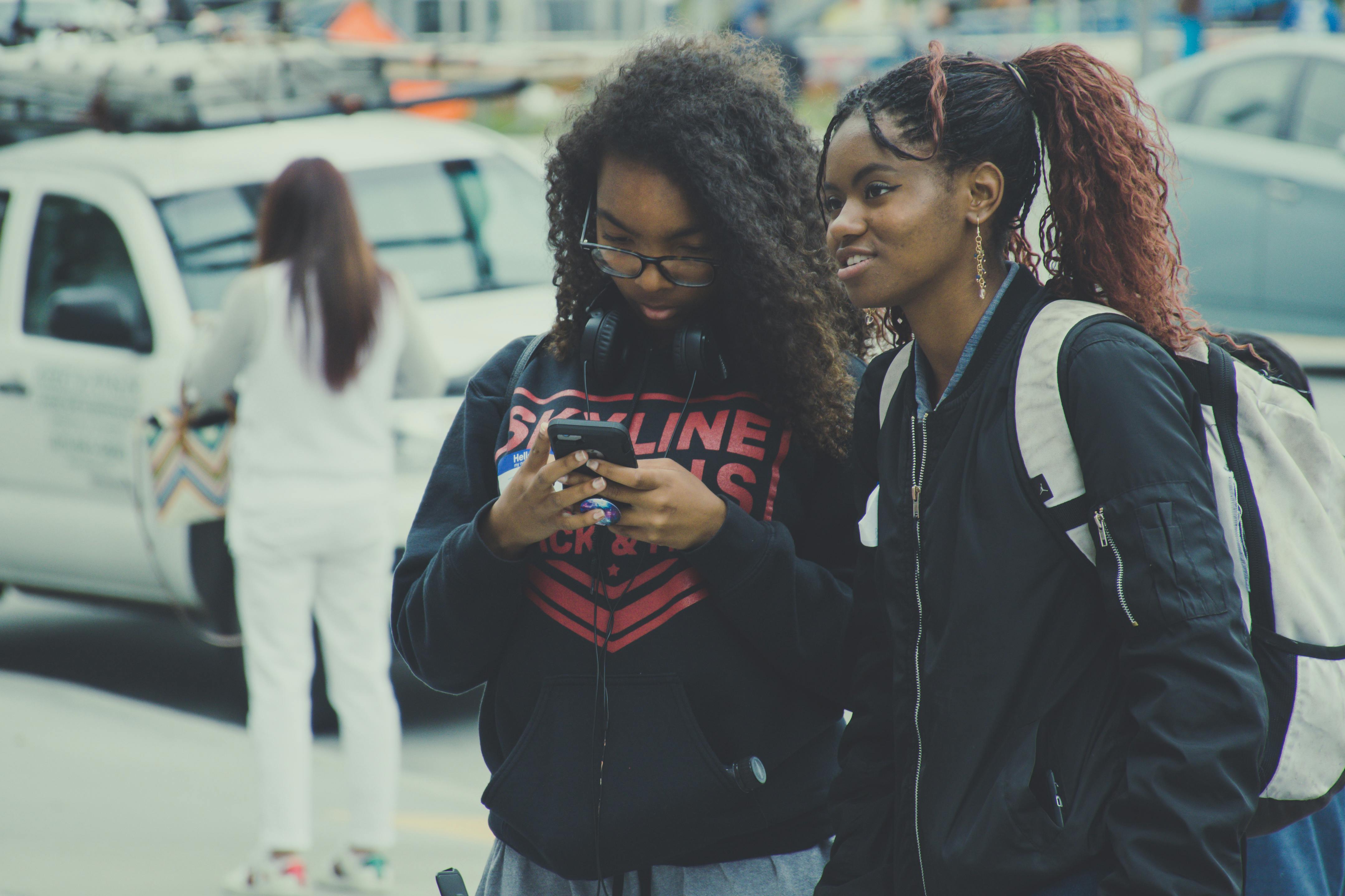 Two girls standing outside together; one girl wears a backpack and the other holds a cell phone