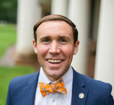 Conor is smiling at the camera wearing a blue jacket, white shirt and very snazzy orange bowtie in front of the columns on the Lawn.